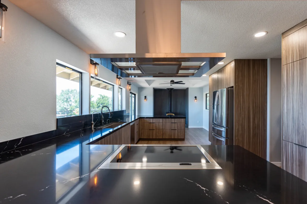 Galley kitchen with black veined stone counters, island cooktop under a stainless hood, walnut-tone cabinets, and ribbon windows