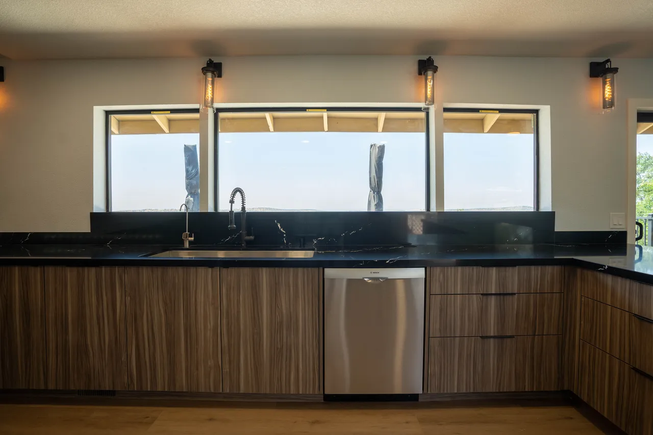 Walnut-tone cabinets with black veined waterfall counter, stainless sink with black spring faucet, and a ribbon window with a hill view
