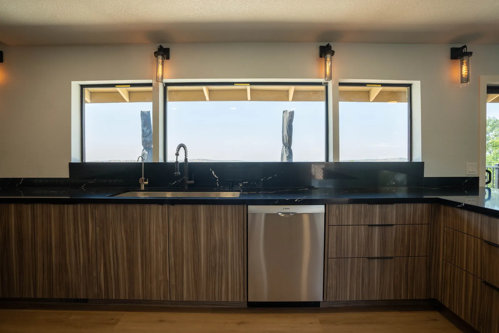 Walnut-tone cabinets with black veined waterfall counter, stainless sink with black spring faucet, and a ribbon window with a hill view