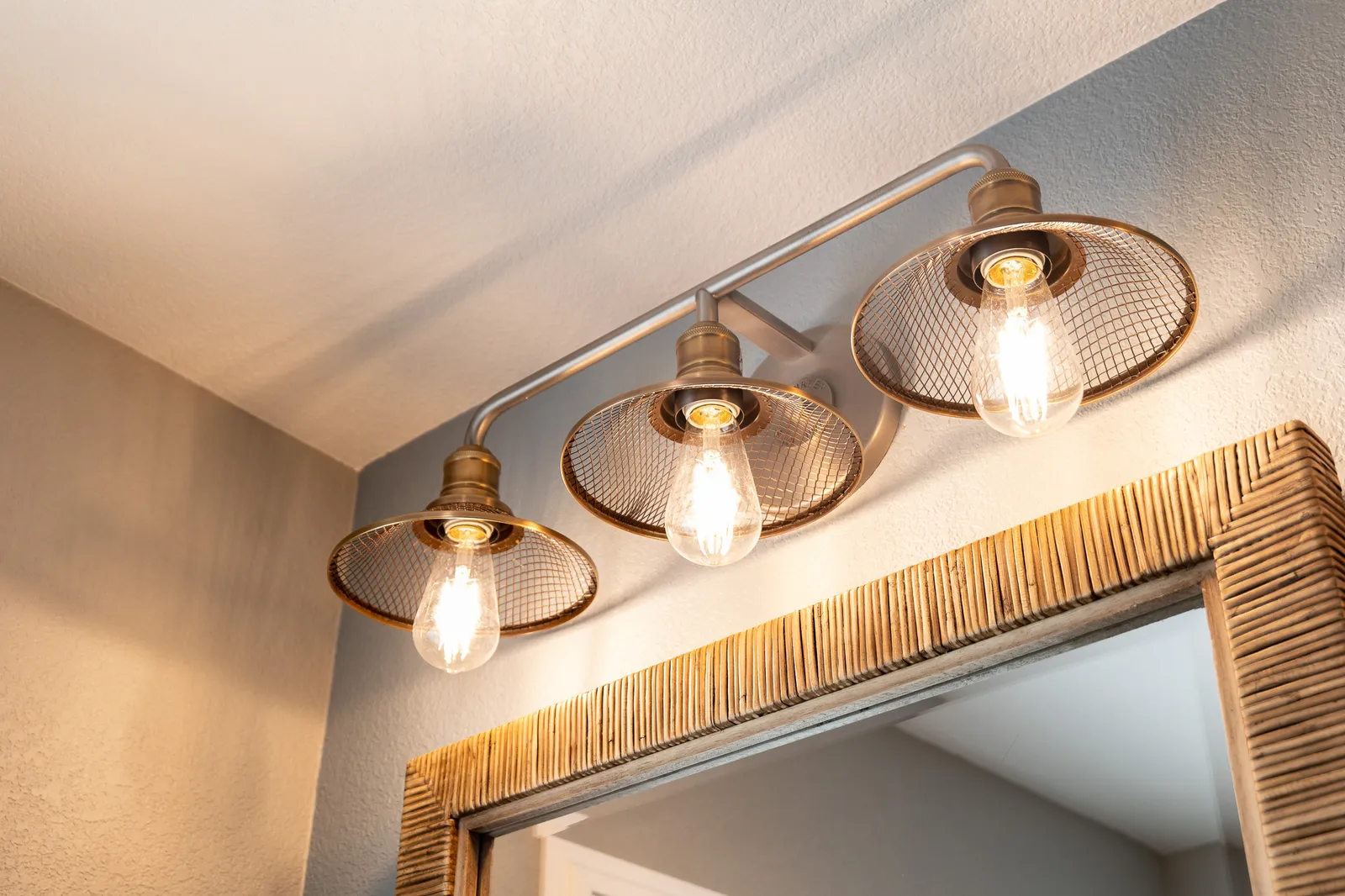 Wire-cage vanity light with exposed bulbs above a woven rattan-framed mirror