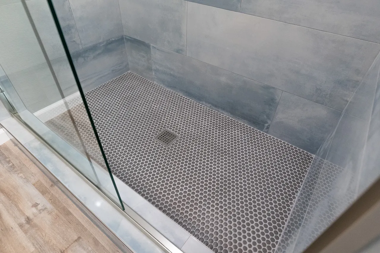 Glass shower with blue-gray large-format wall tile and a gray penny-round mosaic floor with a square drain
