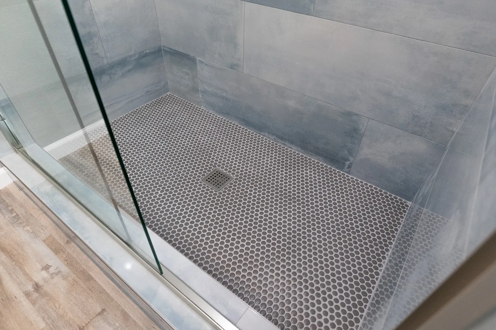 Glass shower with blue-gray large-format wall tile and a gray penny-round mosaic floor with a square drain