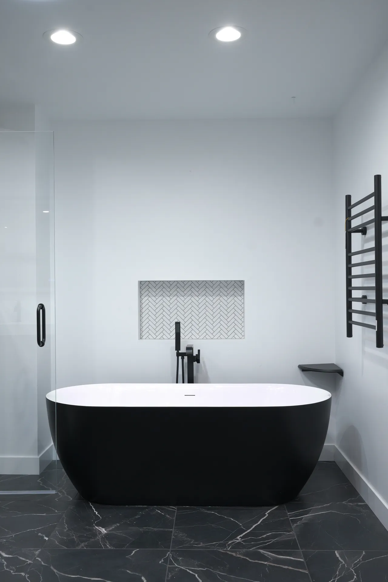 Matte black freestanding tub below a herringbone tile niche, with a black towel warmer and black marble floor