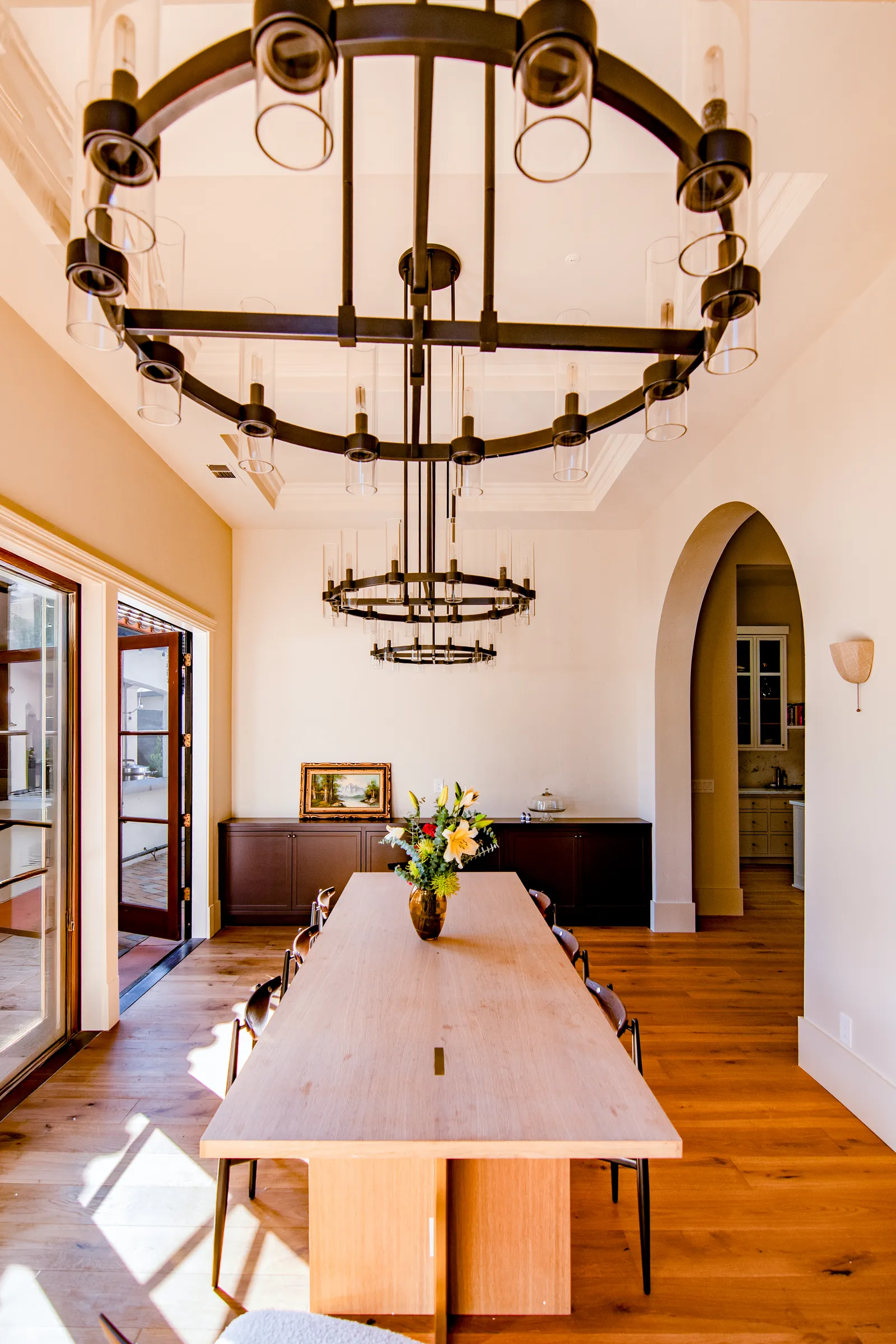 Dining room with tiered iron ring chandeliers over a long wood table, French doors, and an arched opening