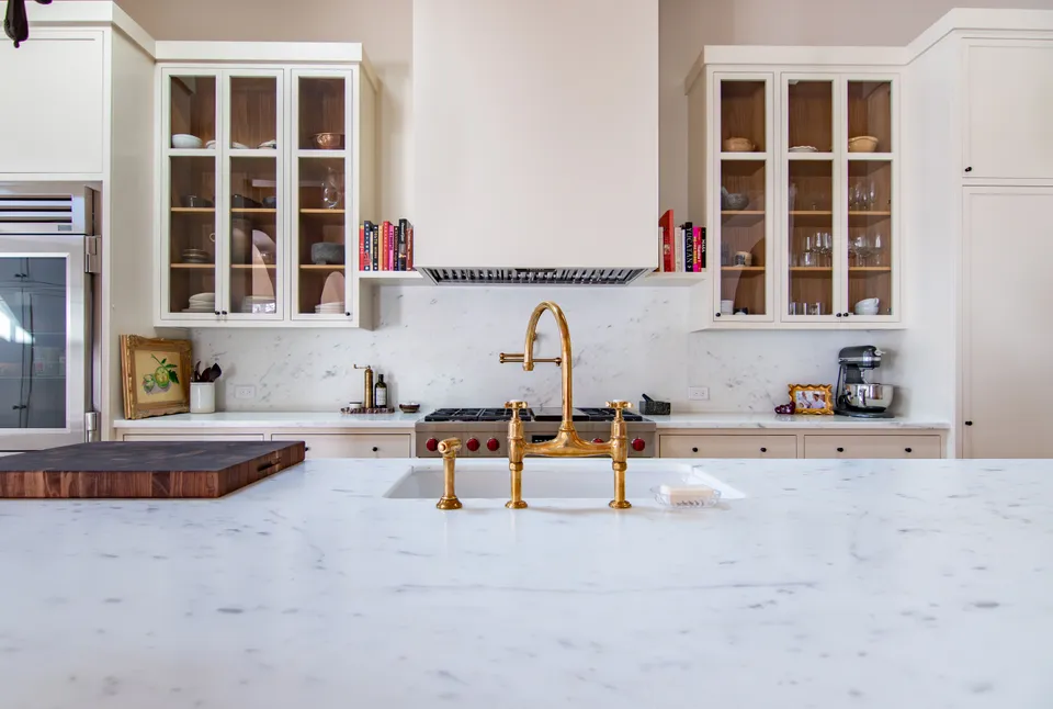 Book-matched Carrara marble island with a brass bridge faucet and glass-front upper cabinets by KHB Construction